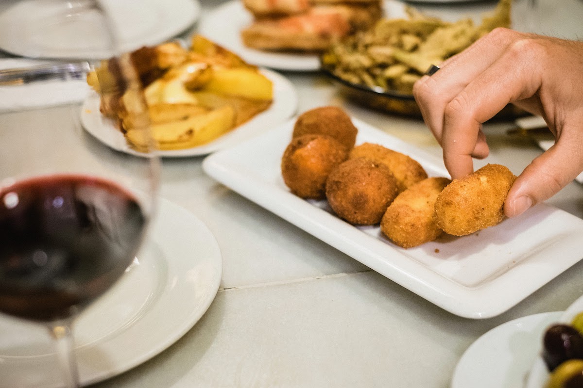 A person's hand taking a fried croquette from a small white dish, with a glass of red wine visible in the foreground.