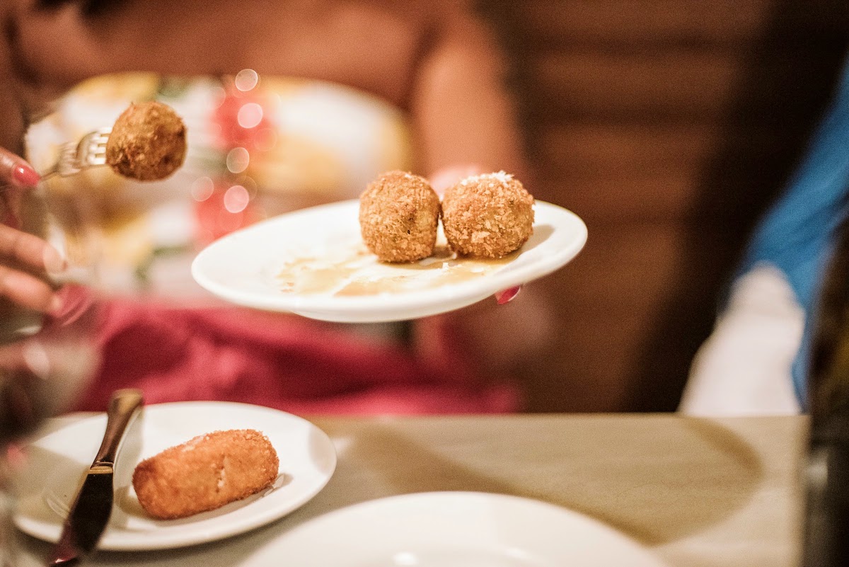 A person using a fork to take a fried croquette off a white plate.