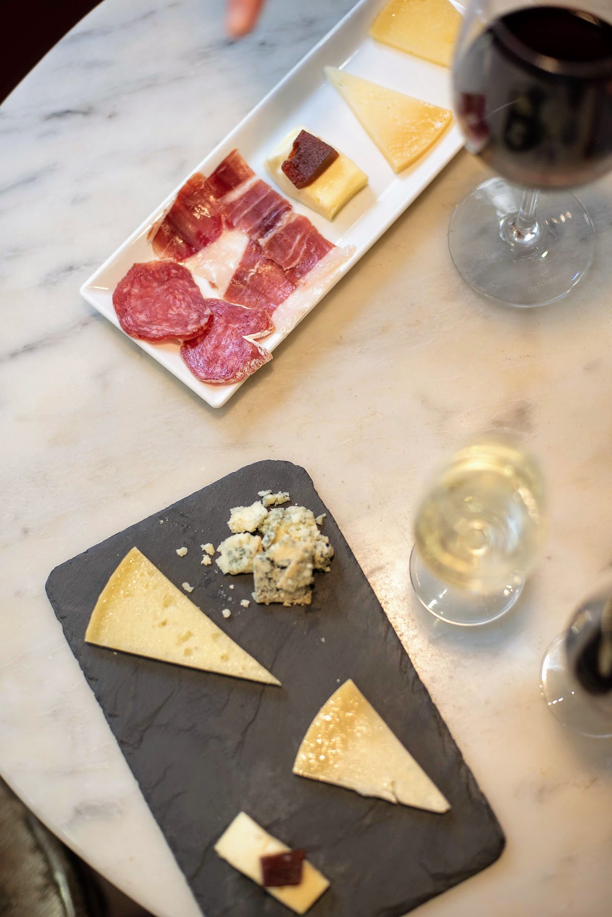 Overhead shot of two small trays of cheeses and cured meats next to a glass of red wine and a smaller glass of pale yellow sherry wine