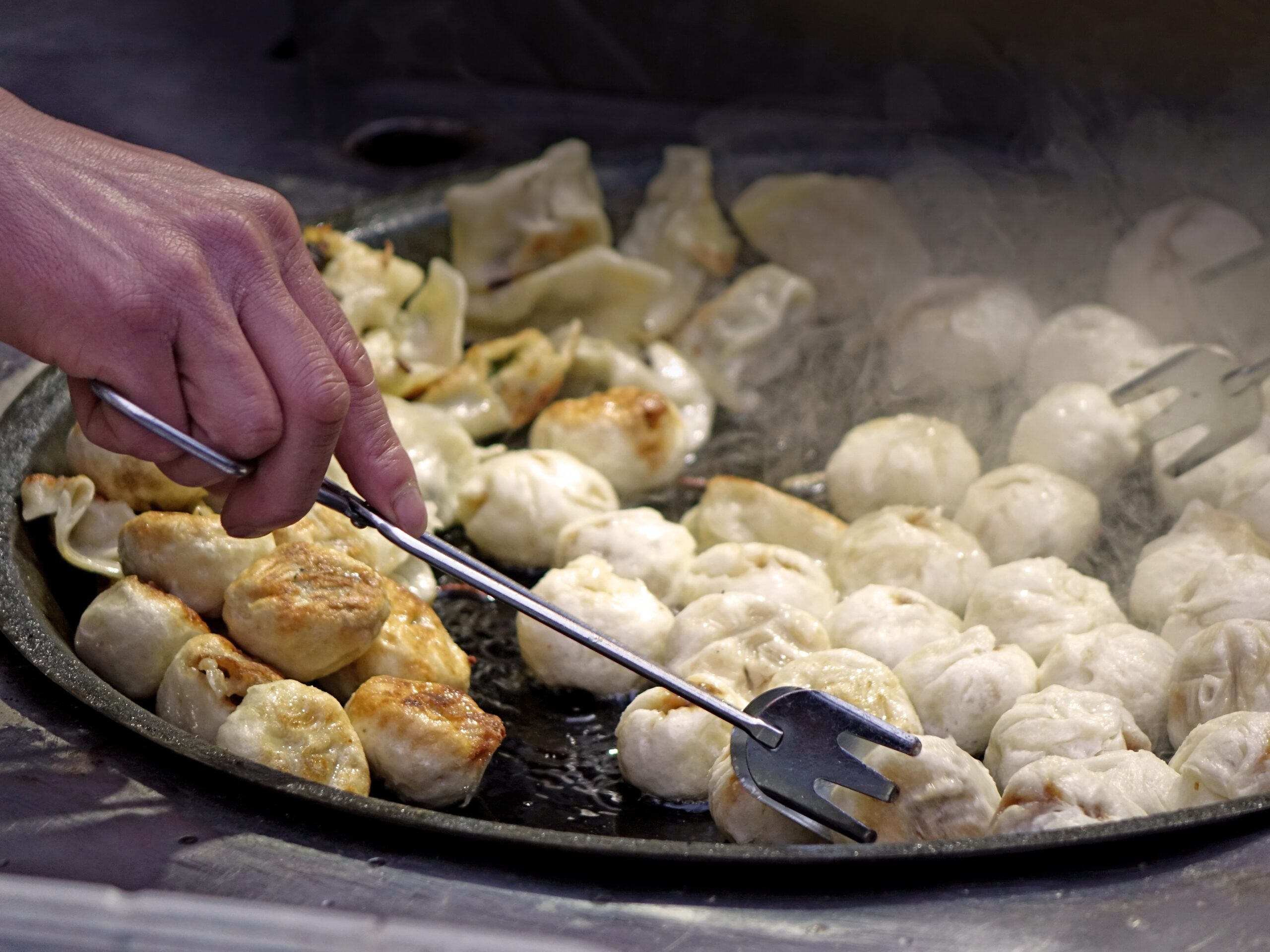 Chinese dumplings being steamed in a large iron skillet with steam and oil