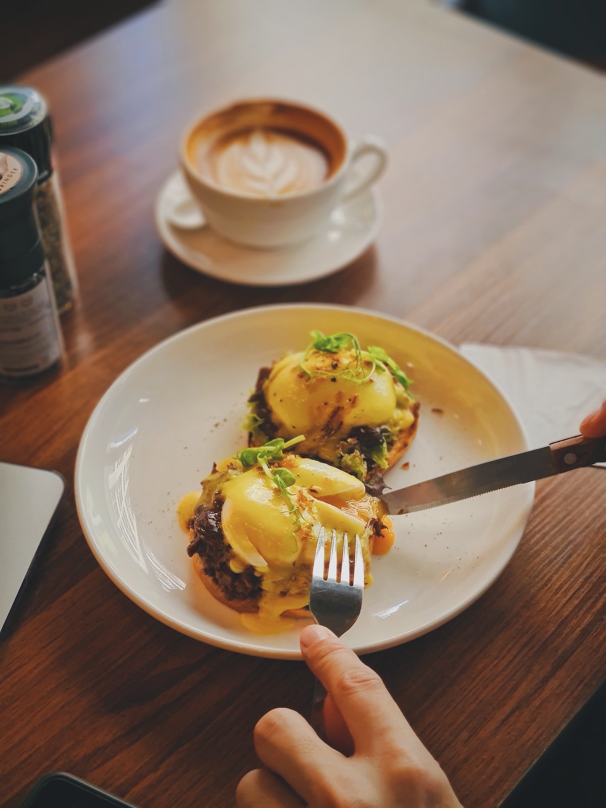 Person cutting into eggs Benedict with a cup of coffee on the table in the background