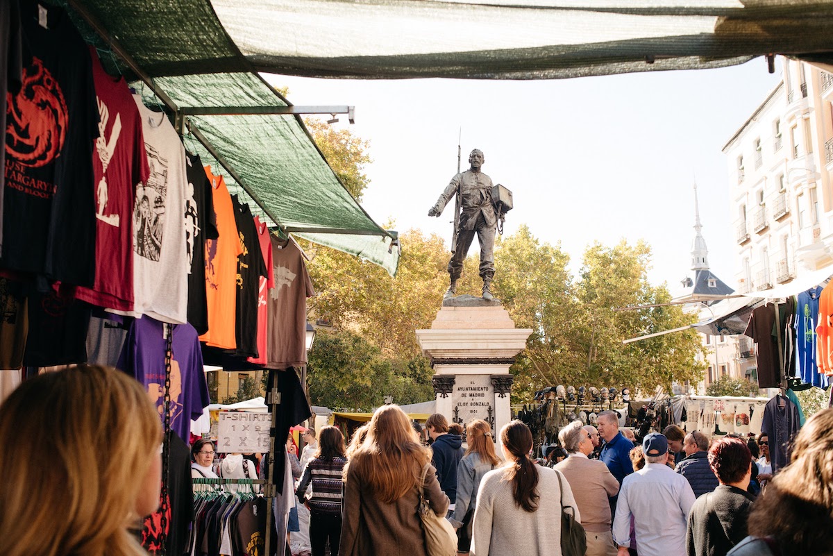 Flea market stalls set up in a busy plaza surrounding a large metal statue of a soldier.