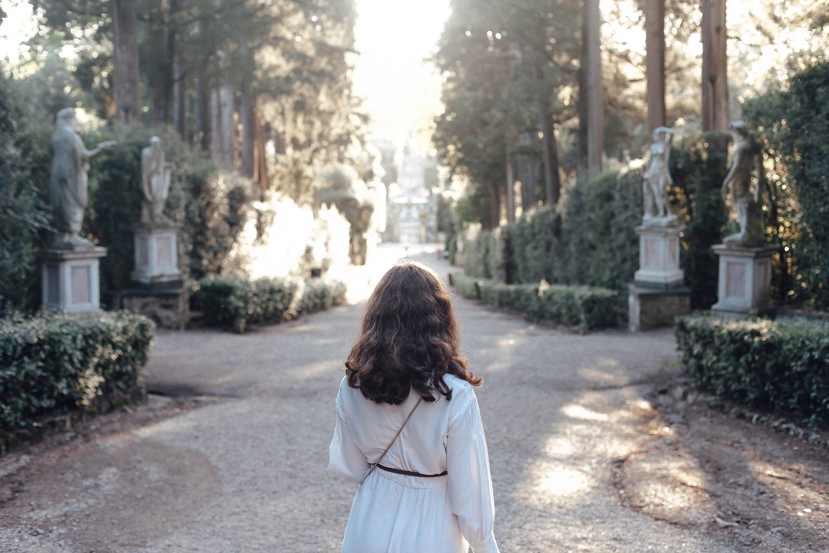 Woman in a white dress walks through a tree-lined boulevard with statues on either side