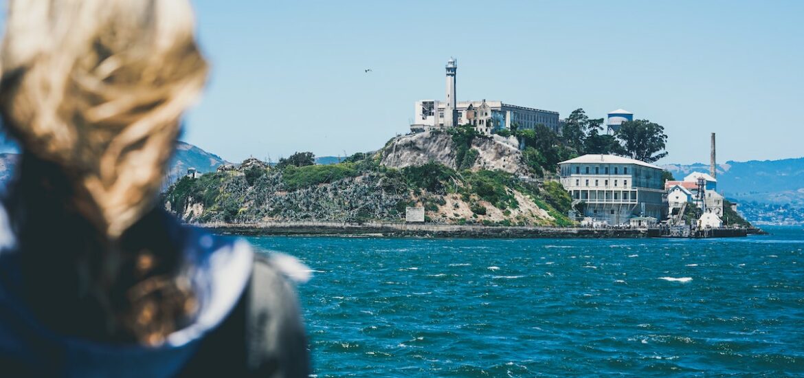 A person with windblown hair in the foreground looks across the Bay to Alcatraz Island, a spooky place to visit in San Francisco