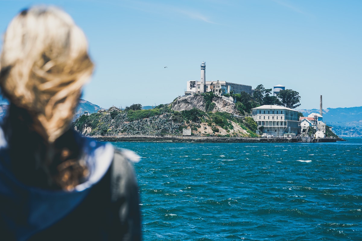 A person with windblown hair in the foreground looks across the Bay to Alcatraz Island, a spooky place to visit in San Francisco