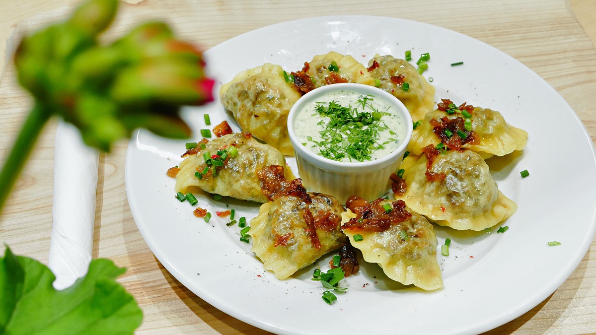 Pierogi or pierogies on a plate with onion, greens, and bacon as well as a yogurt dipping sauce