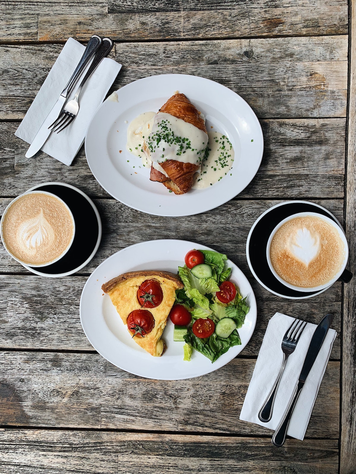 Flat lay of a wooden table with two cappuccinos and a plate with quiche and salad, the other with a croissant