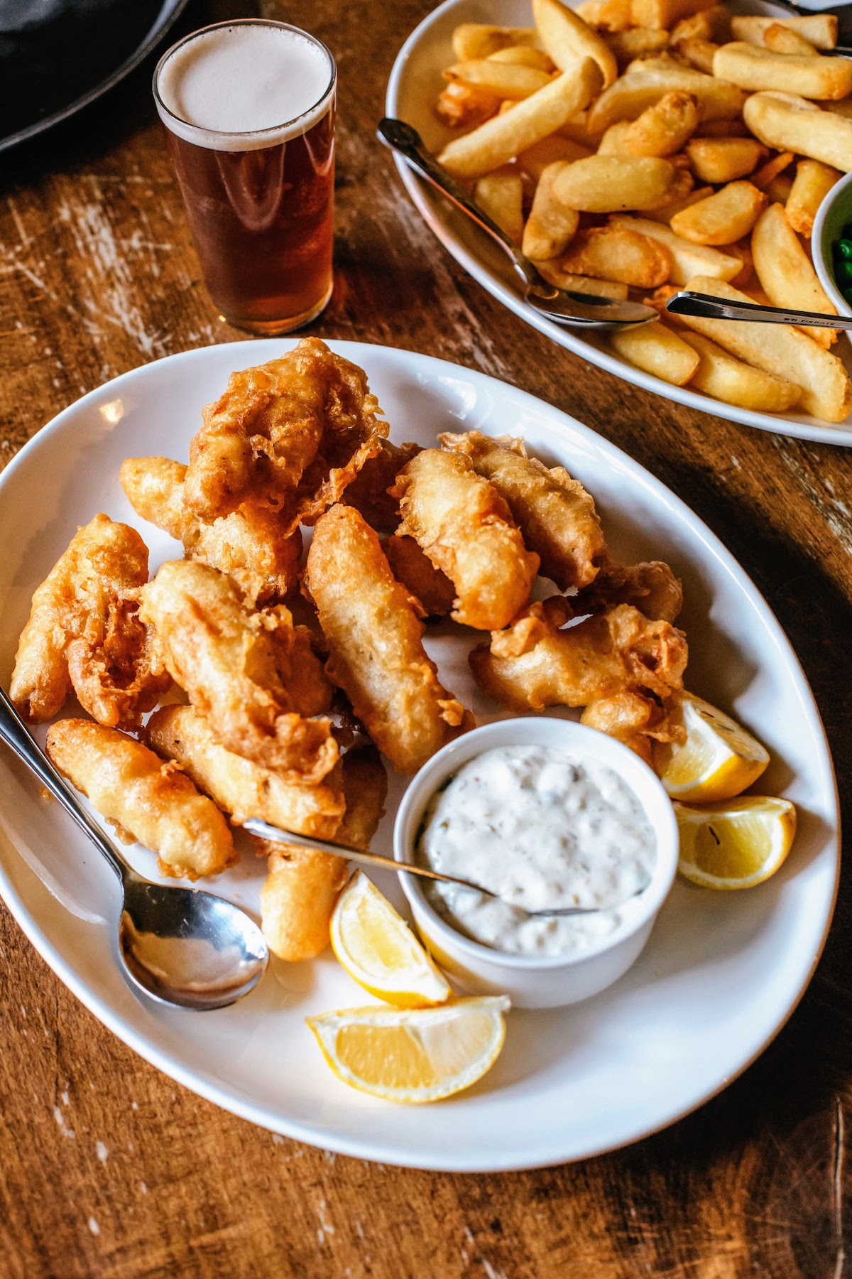 Fried fish pieces served on a white plate with tartar sauce and lemon wedges with a plate of french fries in the background.