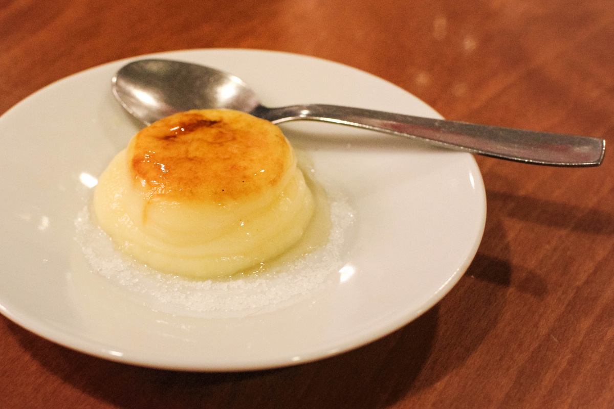 Round piece of flan on a white plate beside a spoon
