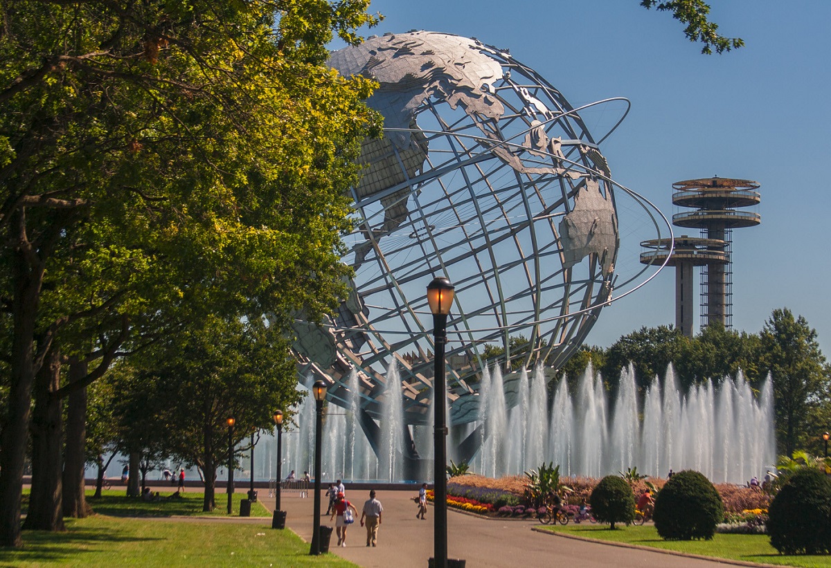 Huge silver sphere shaped like the earth emerges from behind trees and water fountains in a park in new york