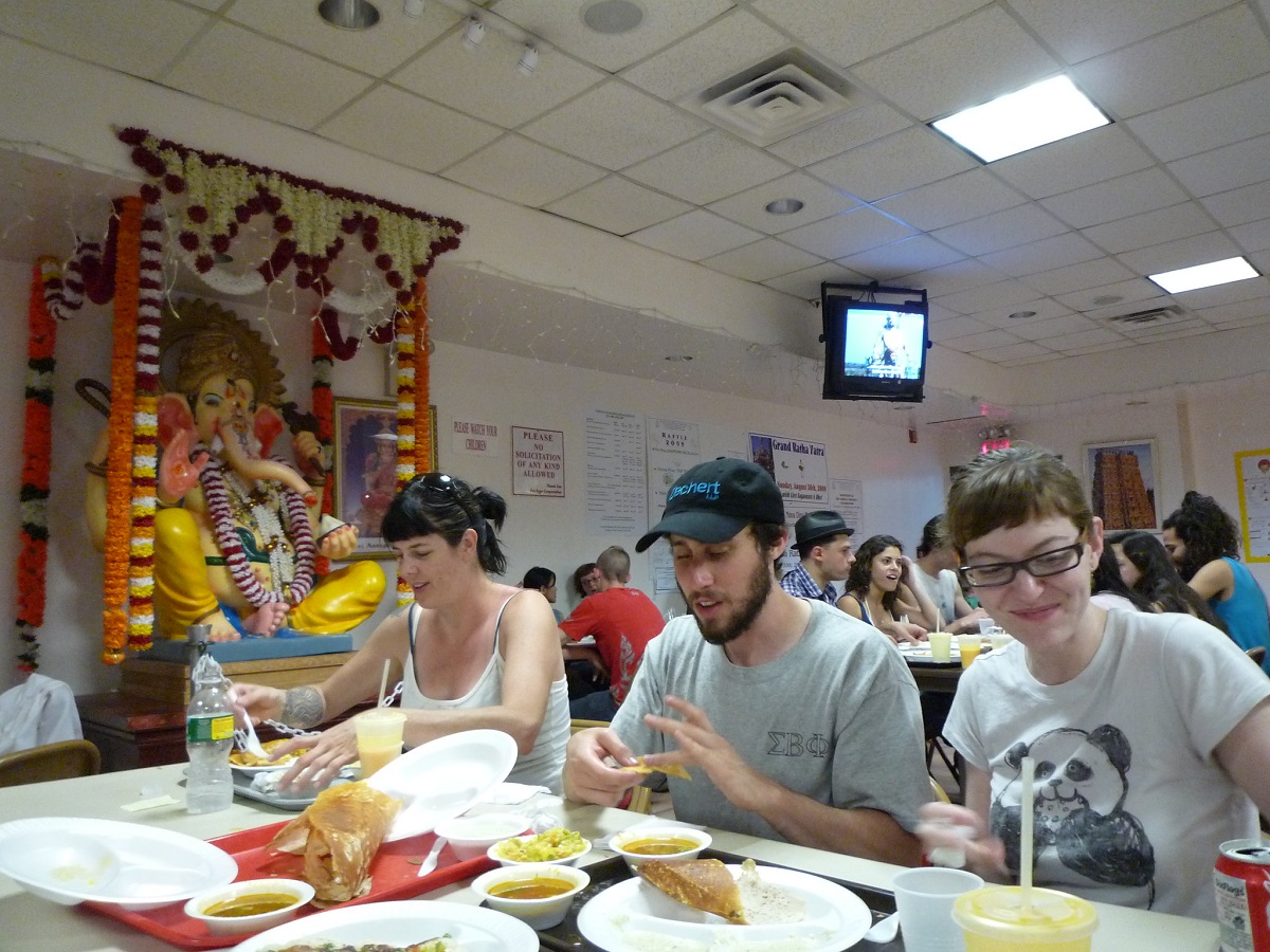 Cafeteria setting inside of a Hindu temple with 4 friends eating food in a no-frills setting