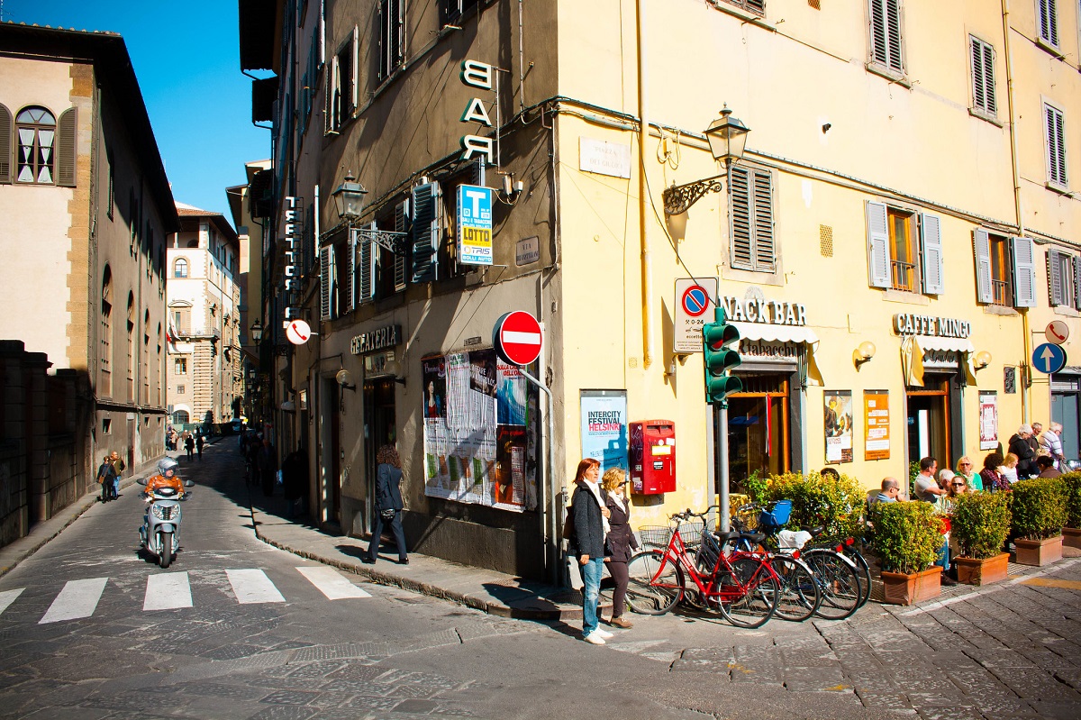 Two women wait outside of a building in the sun with their bikes on a street in Florence