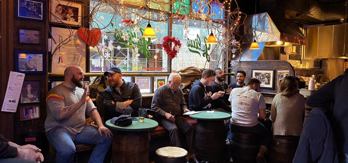 Patrons inside of a bar drink beer around old wooden casks with Christmas lights on the window behind them