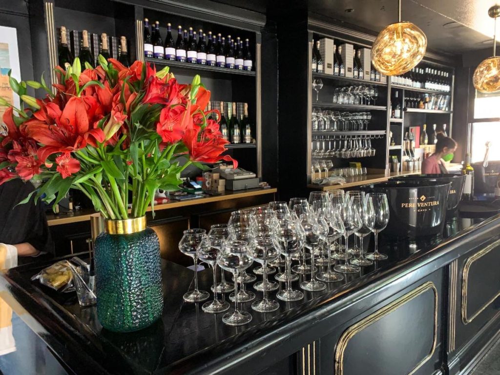 Several wine glasses on a black bar top next to a large vase of red lilies.