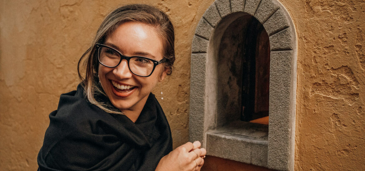 A woman in glasses smiling in front of a small arch-shaped opening in a stone wall