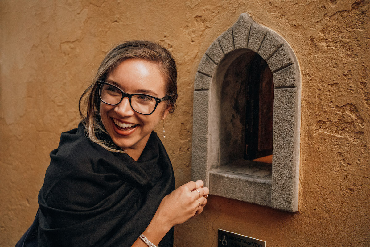 A woman in glasses smiling in front of a small arch-shaped opening in a stone wall