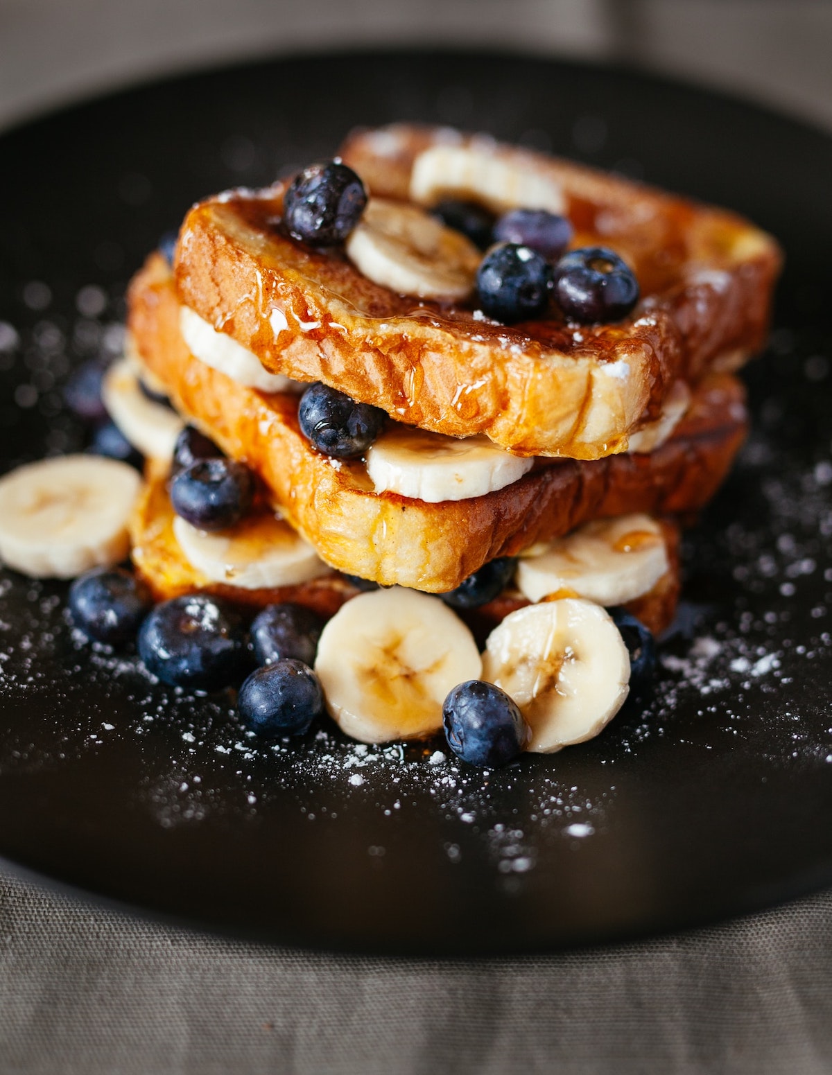 Stack of French toast slices on a black plate with blueberries and banana slices.