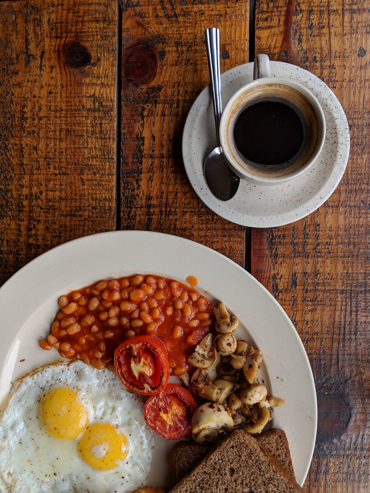 Overhead shot of a full English breakfast including beans, eggs, mushrooms, and tomatoes on a white plate beside a cup of coffee