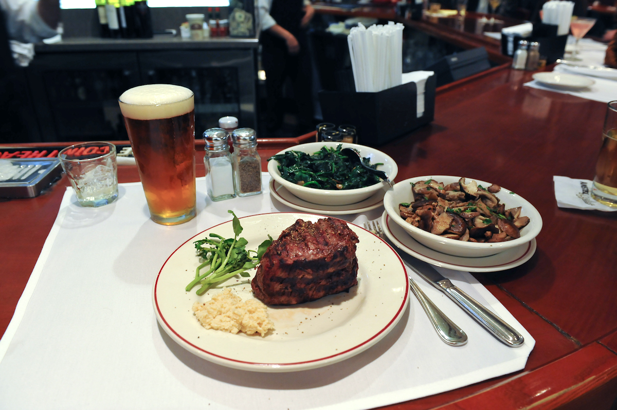 Plate of steak and two dishes of sides on a wooden bar top next to a glass of beer