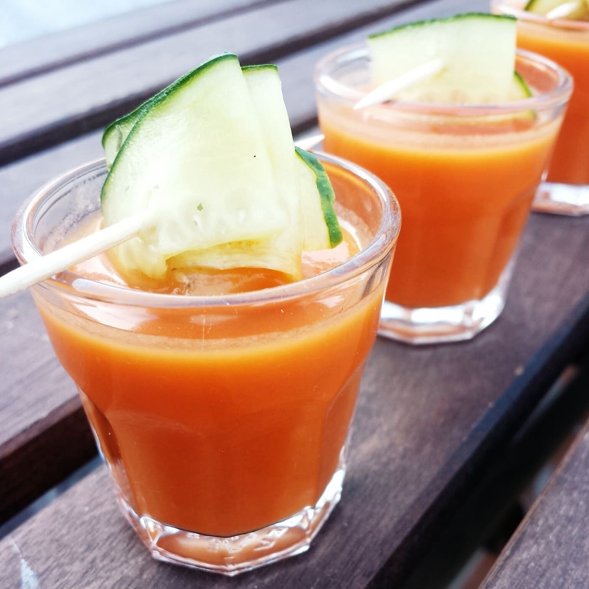 Close up of clear glasses of gazpacho garnished with cucumber on a wooden tabletop.
