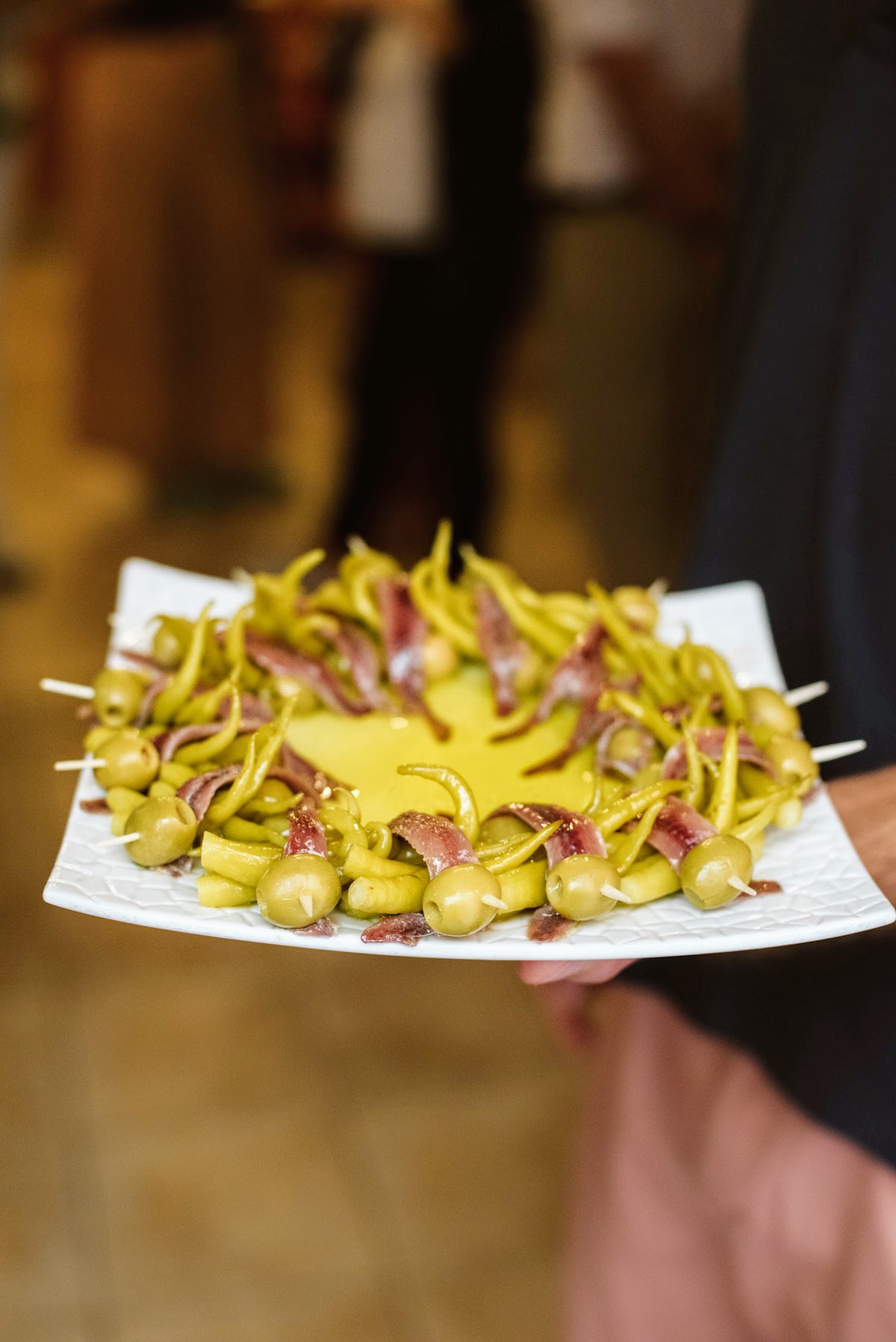 Plate of olive, anchovy, and pepper skewers on a square plate surrounding olive oil