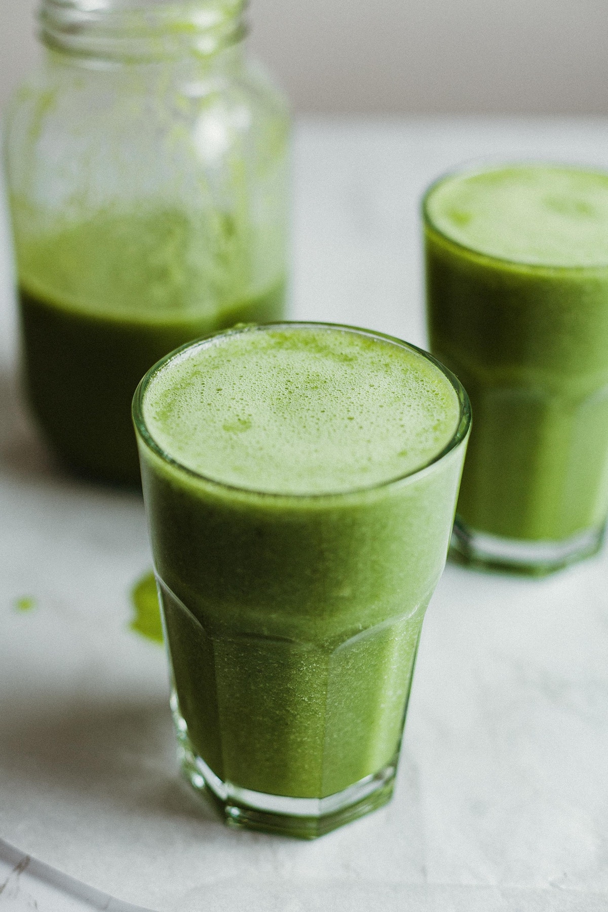 Two glasses of green juice with a larger jar of the juice in the background.