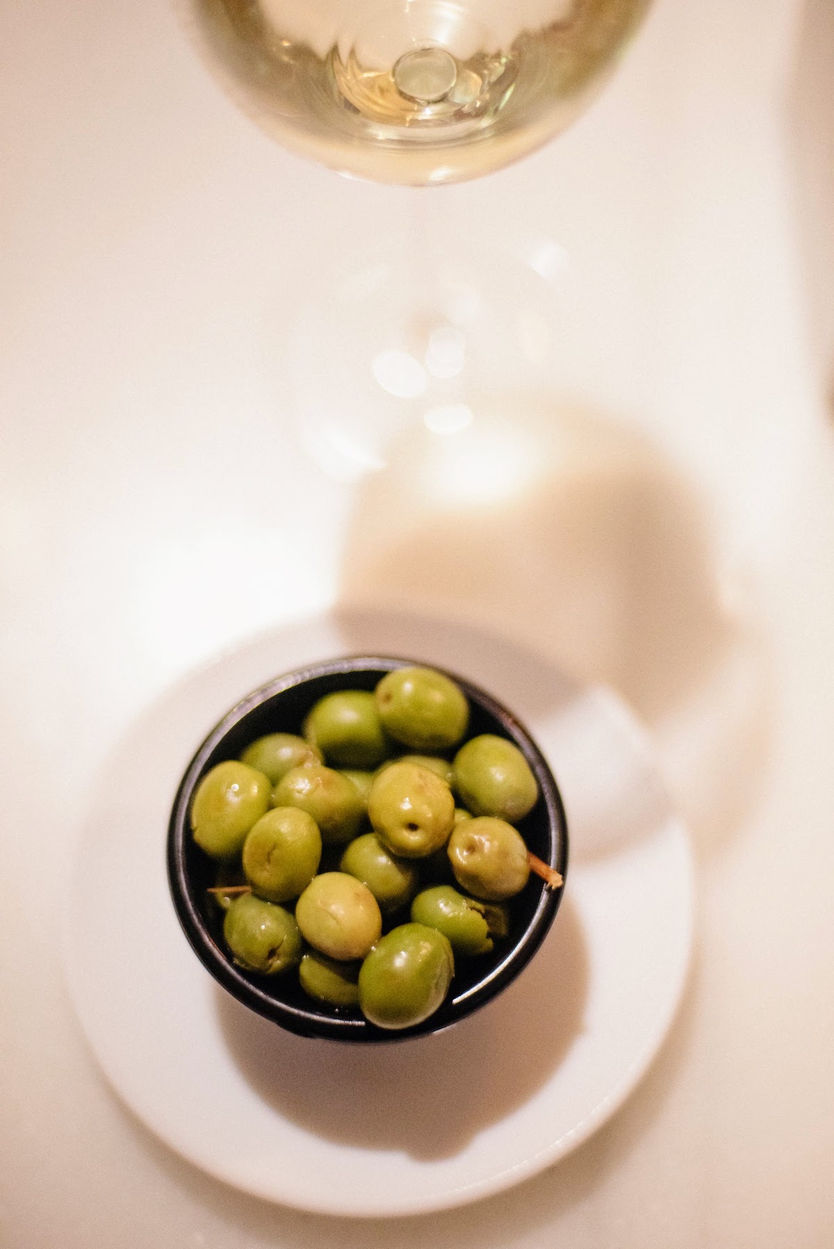 Overhead shot of a small dish of green olives on a white tabletop, with a glass of white wine partially visible behind it.