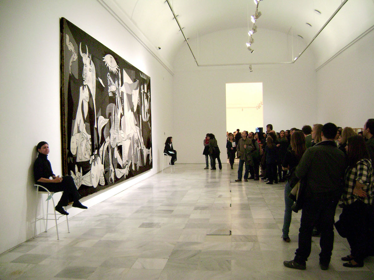 Crowd of people in a white museum room looking at a large black and white painting on the wall.