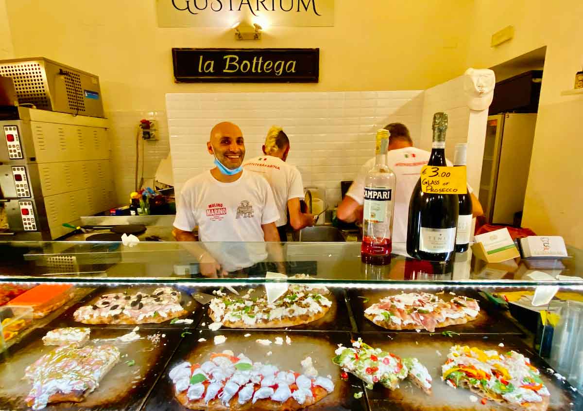 A man smiling behind a counter with several pizzas on display