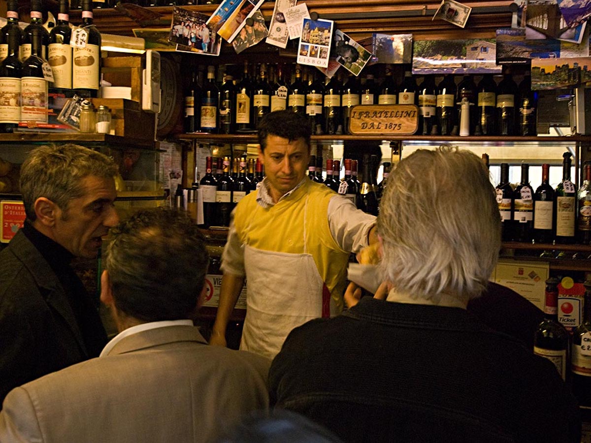 Men standing at a crowded bar in Florence while a bartender serves a panini sandwich.