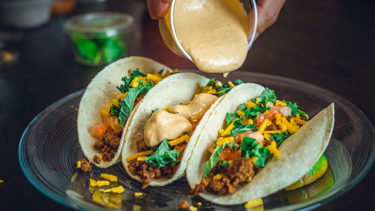 A person pours creamy sauce over three vegan tacos filled with fake meat, lettuce, and tomato
