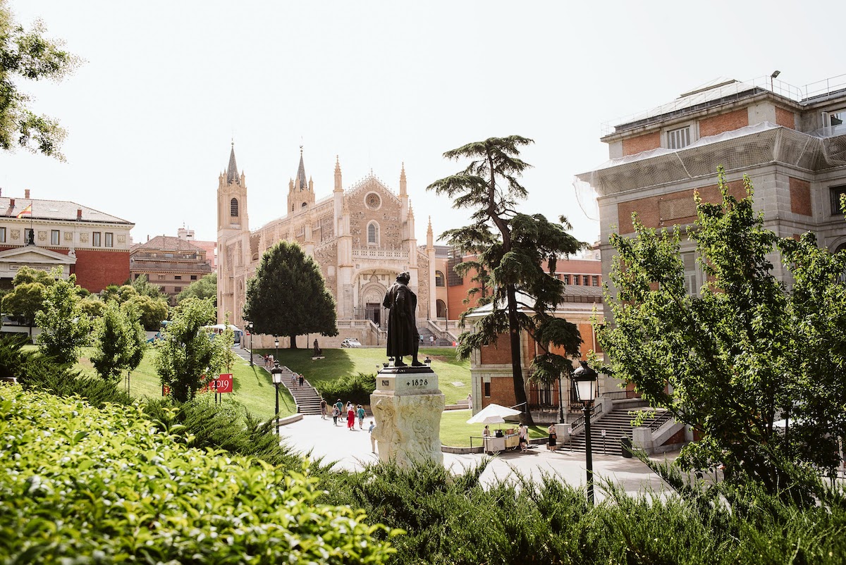 Scene from Madrid depicting a large white church, the Prado museum, and a statue of a man, all surrounded by greenery.
