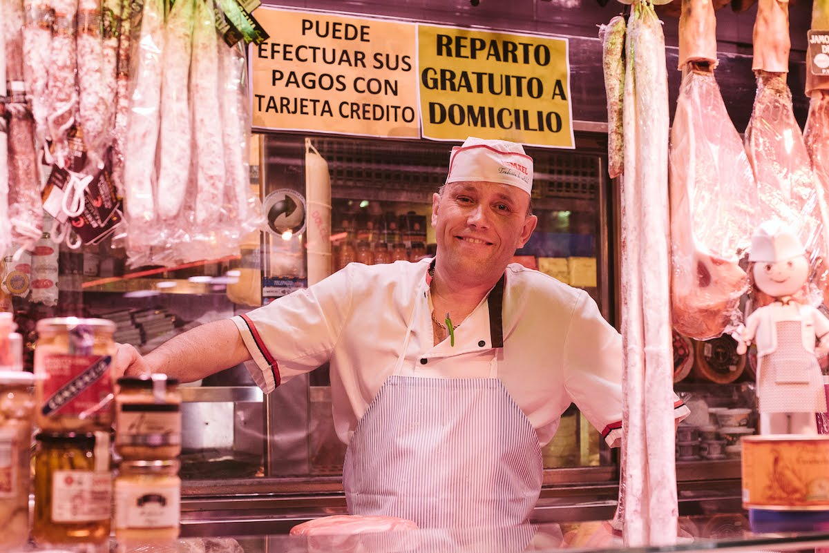 A smiling man at a ham and charcuterie stall at a food market.