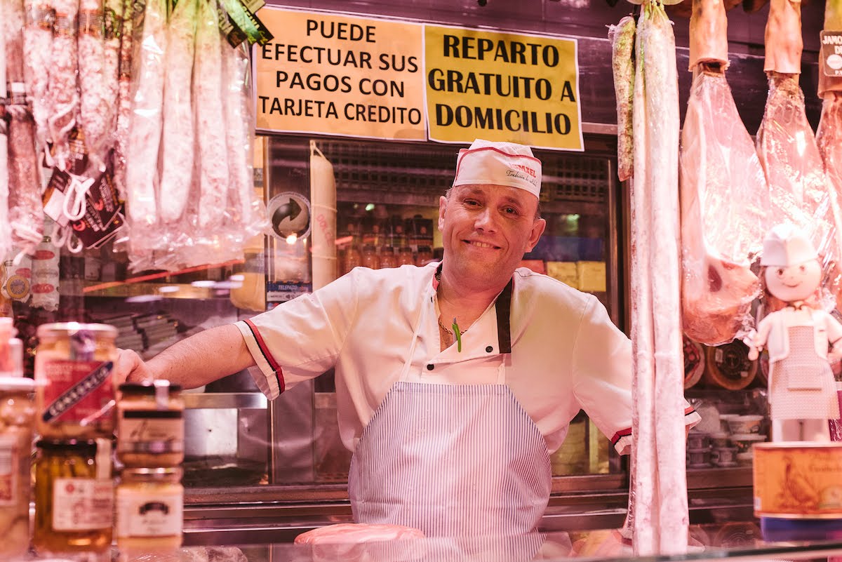 A smiling man at a ham and charcuterie stall at a food market. 