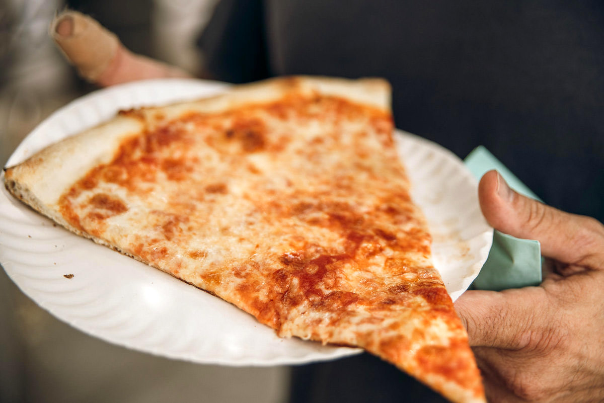 Large slice of New York style pizza on a white paper plate.