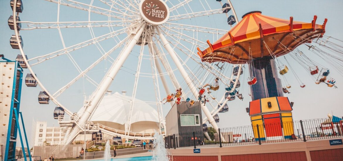 wide shot of Navy Pier in Chicago, with a large white Ferris Wheel in the background, a red and yellow swing ride to the right, and a fountain in the foreground