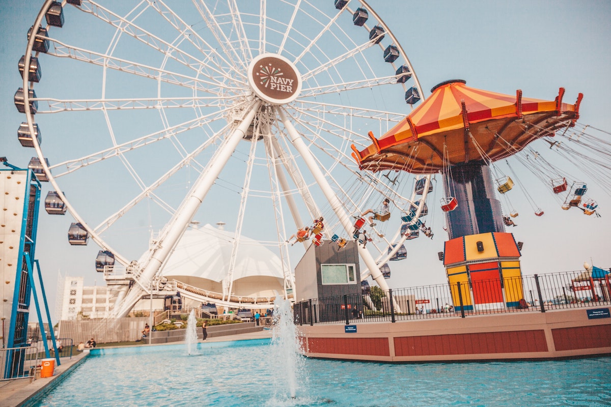 wide shot of Navy Pier in Chicago, with a large white Ferris Wheel in the background, a red and yellow swing ride to the right, and a fountain in the foreground