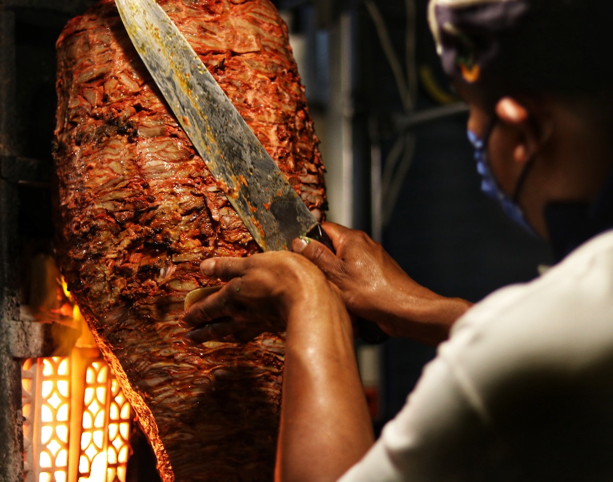 A man using a large knife to cut kebab meat off a vertical spit