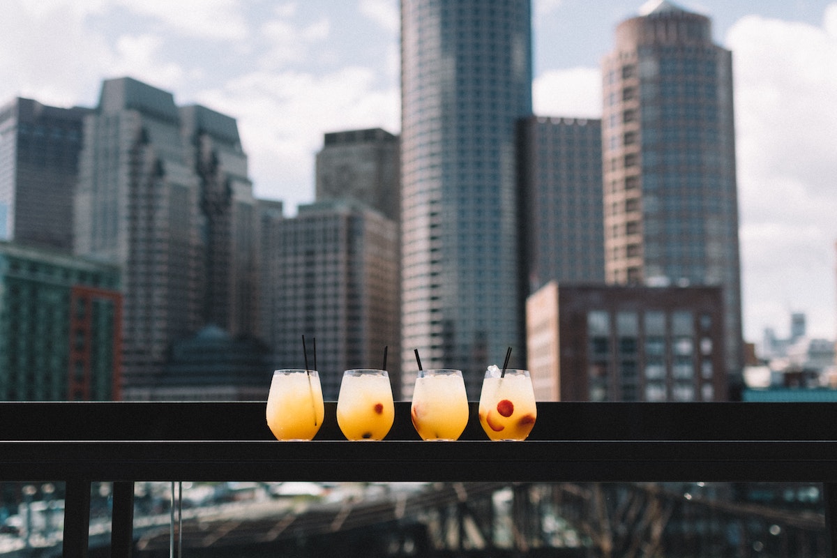 Four brightly colored cocktails sit atop a guardrail on a rooftop cocktail bar in Boston with the city skyline in the background.