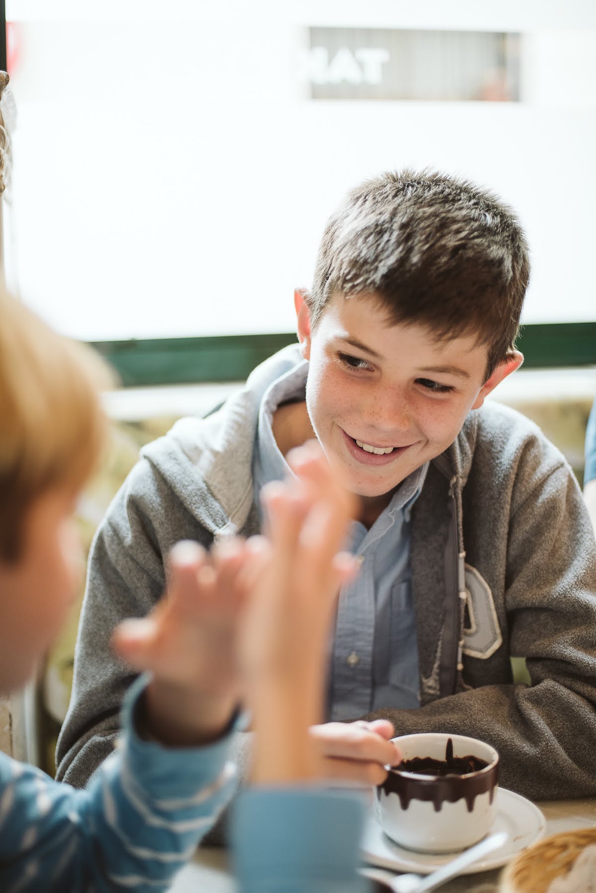 A boy smiling while drinking a mug of hot chocolate while looking at another boy seated at the same table just out of focus