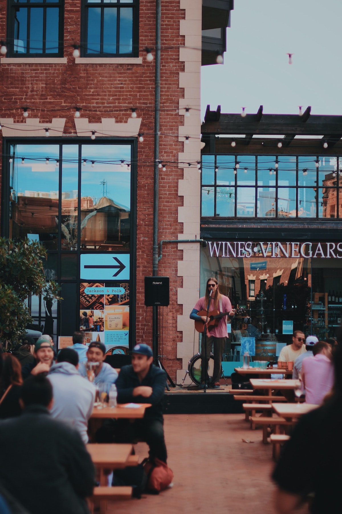 An outdoor courtyard in San Francisco where people are sitting at picnic tables, with someone playing guitar and singing into a microphone