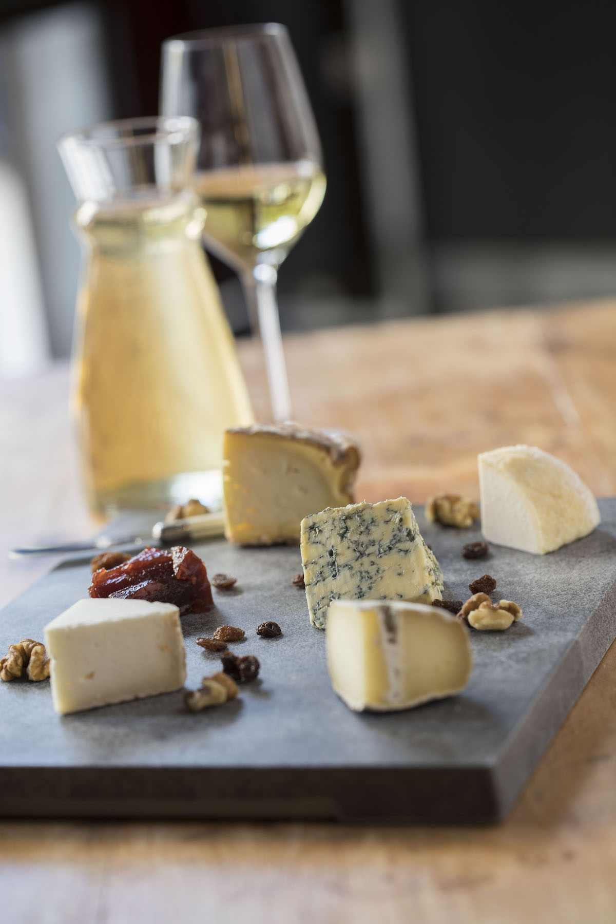 Small chunks of cheese arranged with nuts on a dark gray board, with a glass of white wine in the background.