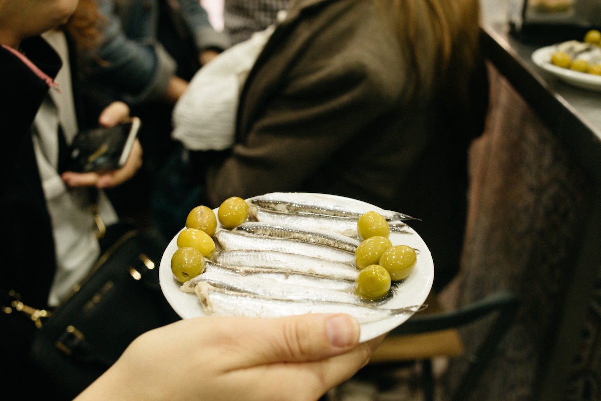 Person's hand holding a plate of anchovies and green olives.