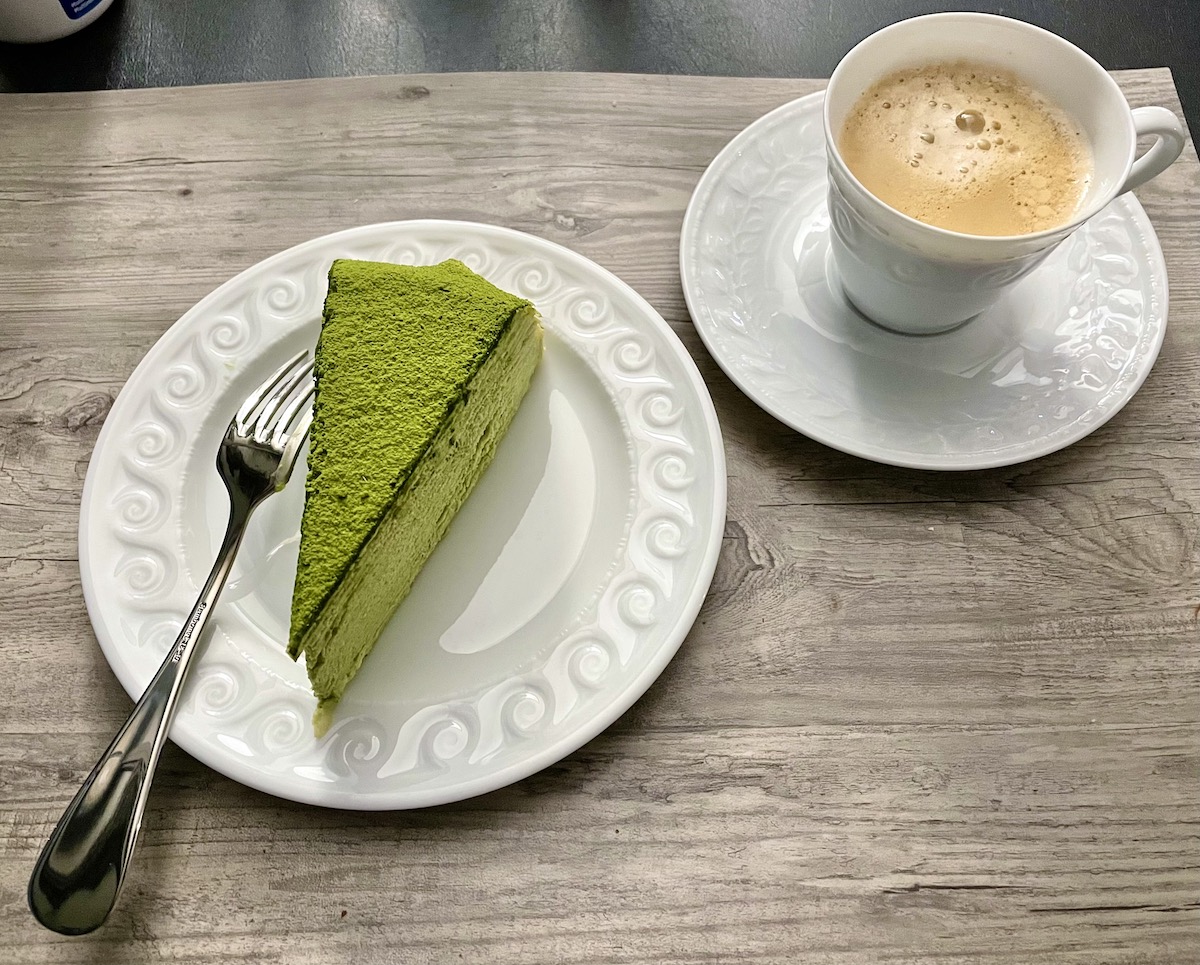 Slice of green cake on a white plate beside a cup of coffee on a wooden tabletop