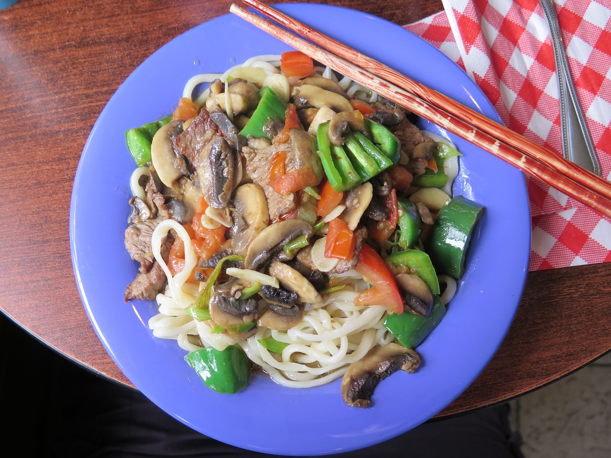 Overhead shot of an Asian noodle dish with mushrooms and peppers on a blue plate with a pair of light red chopsticks