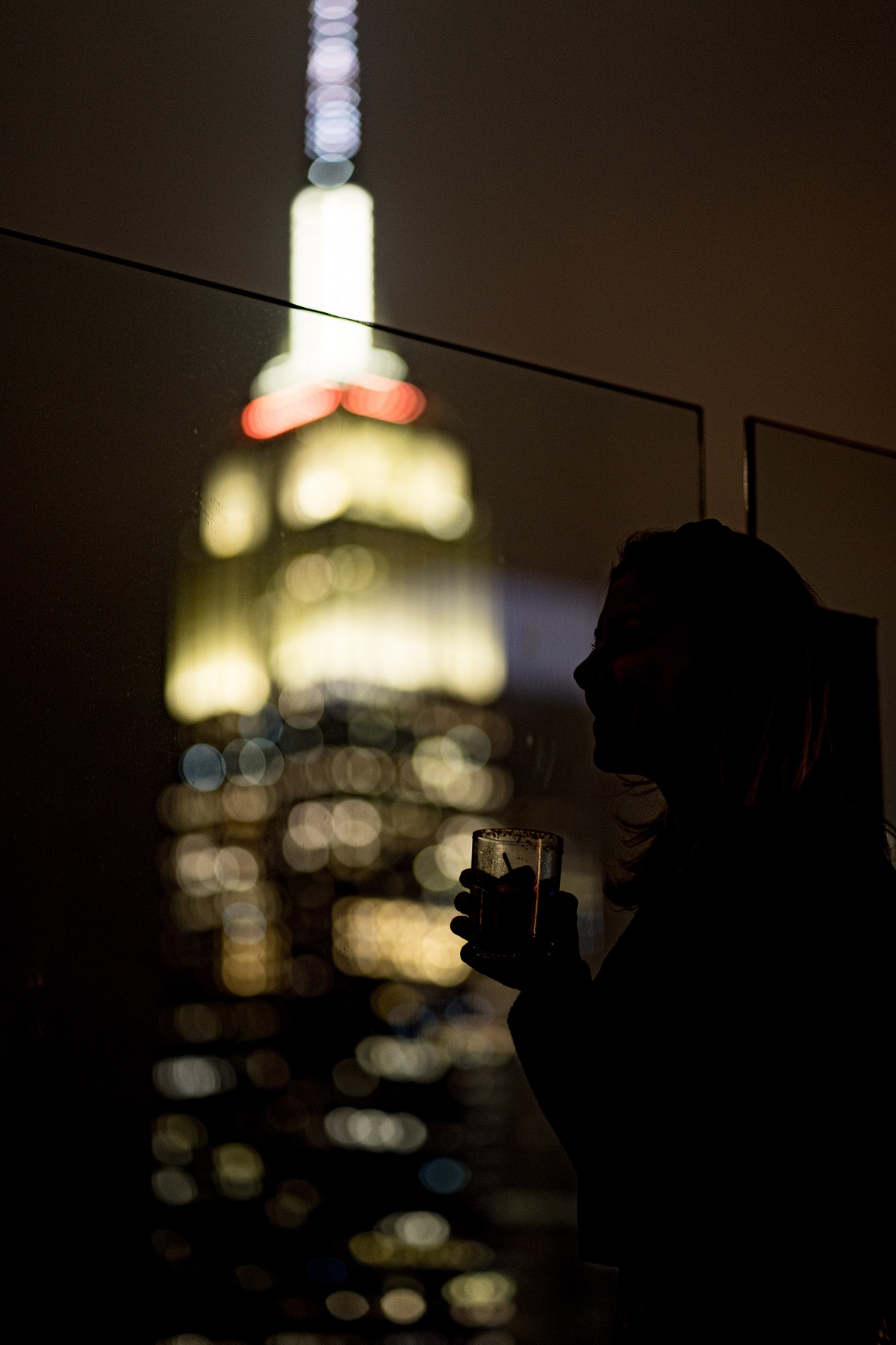 A woman drinks a cocktail at night with the lights of manhattan lighting up the background of the dark photo.