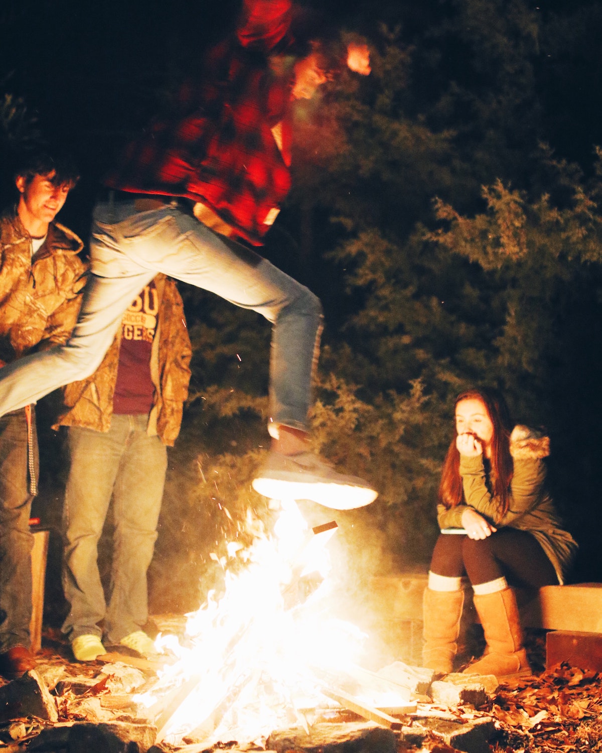 Man in a plaid shirt and jeans leaping over a bonfire as three other people look on.
