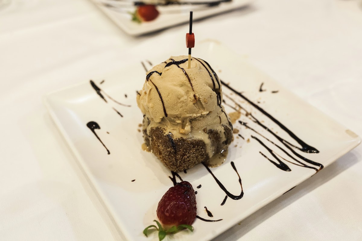 A small square of fried custard topped with a scoop of light brown ice cream, served on a white square plate garnished with chocolate syrup and strawberries.