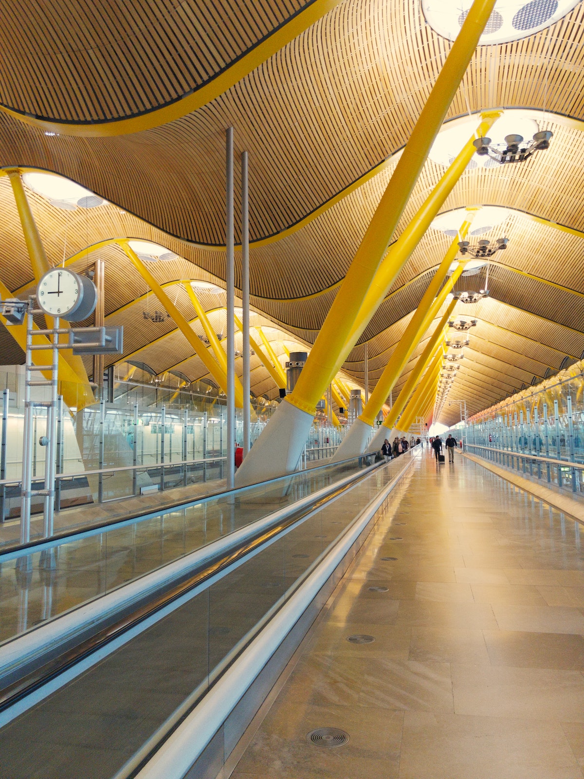 Interior of an airport with a moving walkway and modern ondulated ceiling held up by yellow beams.