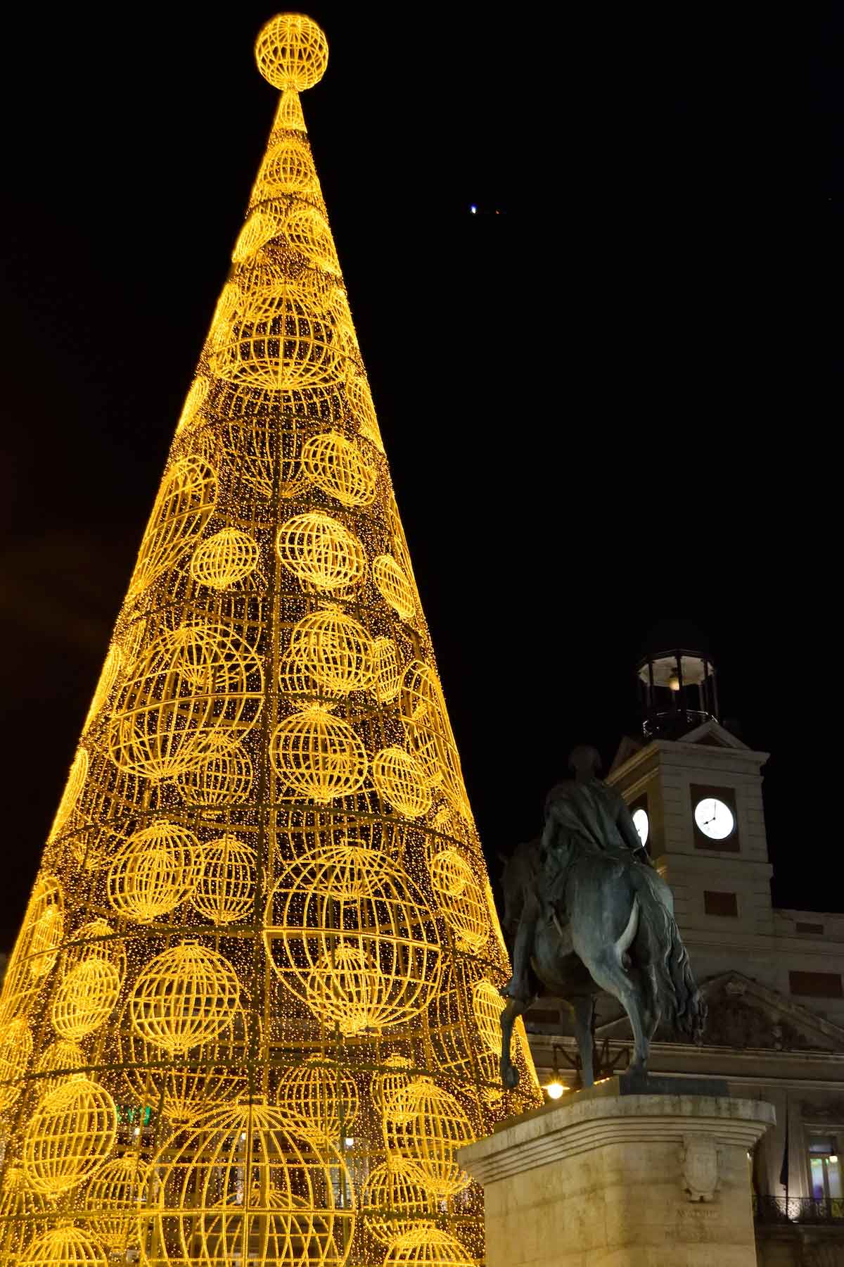 Large outdoor Christmas tree decorated with yellow lights illuminated at night, with a building with a clock tower visible in the background.